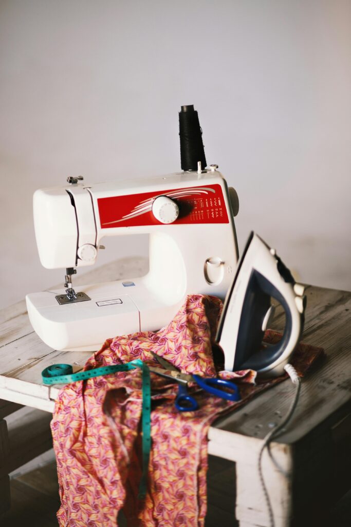 Close-up of a sewing setup with machine, iron, fabric, and tools on a wooden table.